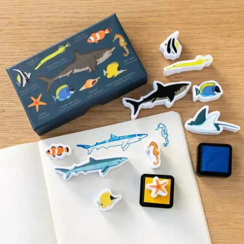 A set of colorful ocean-themed mini stamps placed on a notebook, with a variety of sea creatures such as sharks and fish, alongside two small square ink pads in blue and gray.