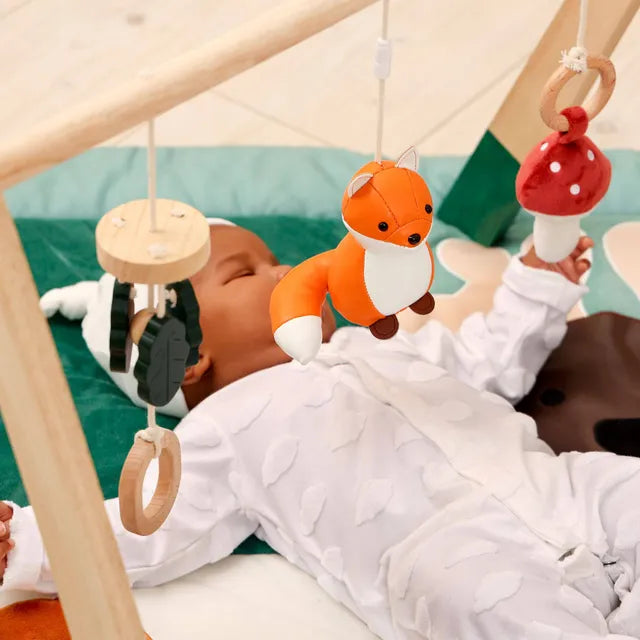 A baby wearing a white patterned onesie lies on their back under a wooden baby gym, reaching for a hanging plush fox toy. Other hanging toys include a wooden ring and a red-and-white mushroom.