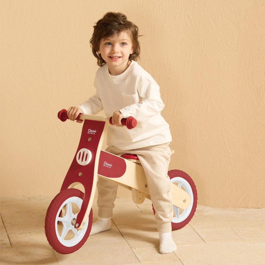 Child riding a red wooden balance bike and beige balance bike against a beige wall.