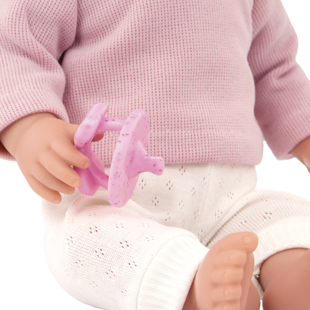 Close-up of a doll wearing a pink sweater and white pants, holding a pink dummy.