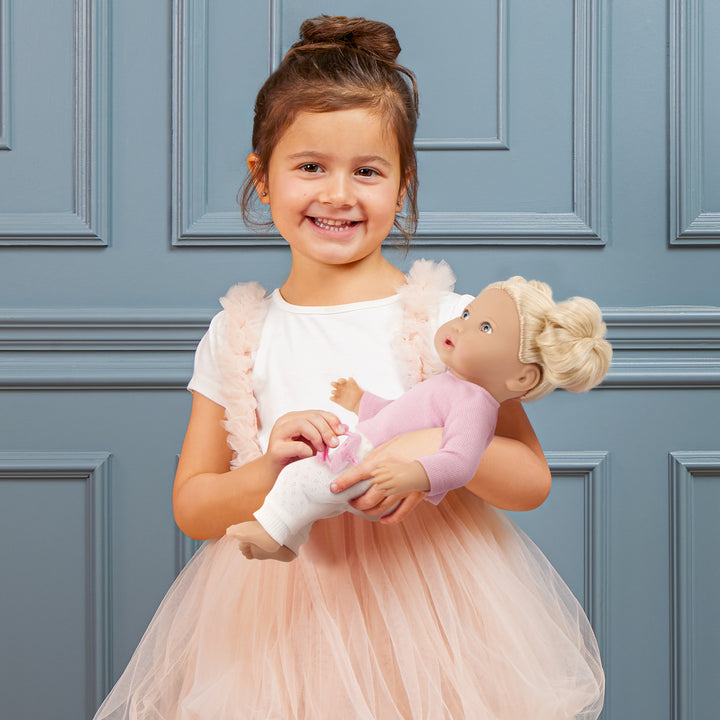 Young girl in a pink dress holding a lullababy doll against a blue paneled wall
