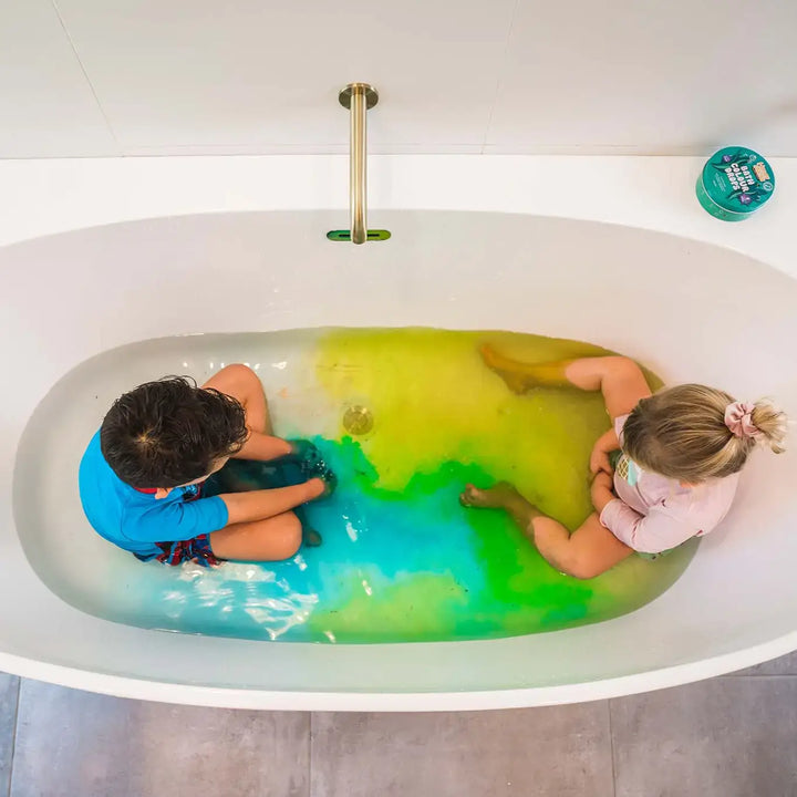 Two children in a bathtub filled with green water, with a visible brand logo on the wall using bath drops