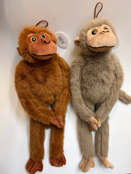 Two plush monkey hanging toys displayed side by side against a plain white wall. The left toy is medium brown with darker brown feet and hands, and the right toy is light gray-brown with beige hands and feet.