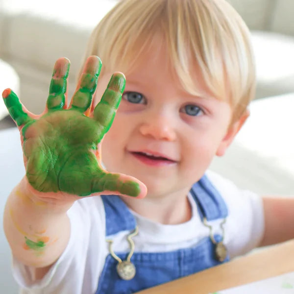 Child with green paint on hand, wearing blue overalls indoors