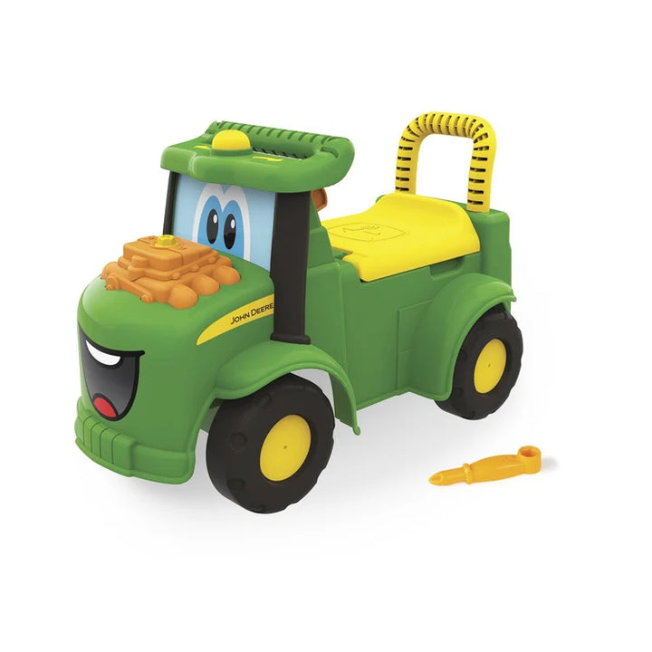 A green and yellow John Deere toy tractor ride-on with a cartoon face, a small pile of gold coins on the hood, and a small yellow toy tool placed beside it on a white background.