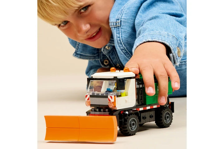 A child wearing a blue denim jacket leans over a toy construction vehicle made of building blocks, with a small orange blade in front on a plain light background.
