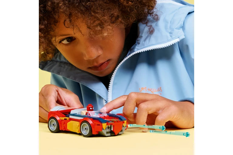 A child wearing a light blue zip-up jacket leans over a small red and yellow toy car with a clear windshield, using their fingers to adjust it on a pale yellow surface.