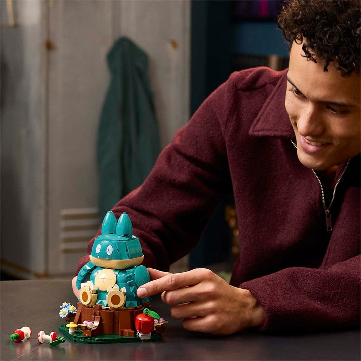 A person in a burgundy fleece jacket smiles while pointing at a small assembled toy structure made of blue, brown, green, and beige building blocks on a table in an indoor setting.