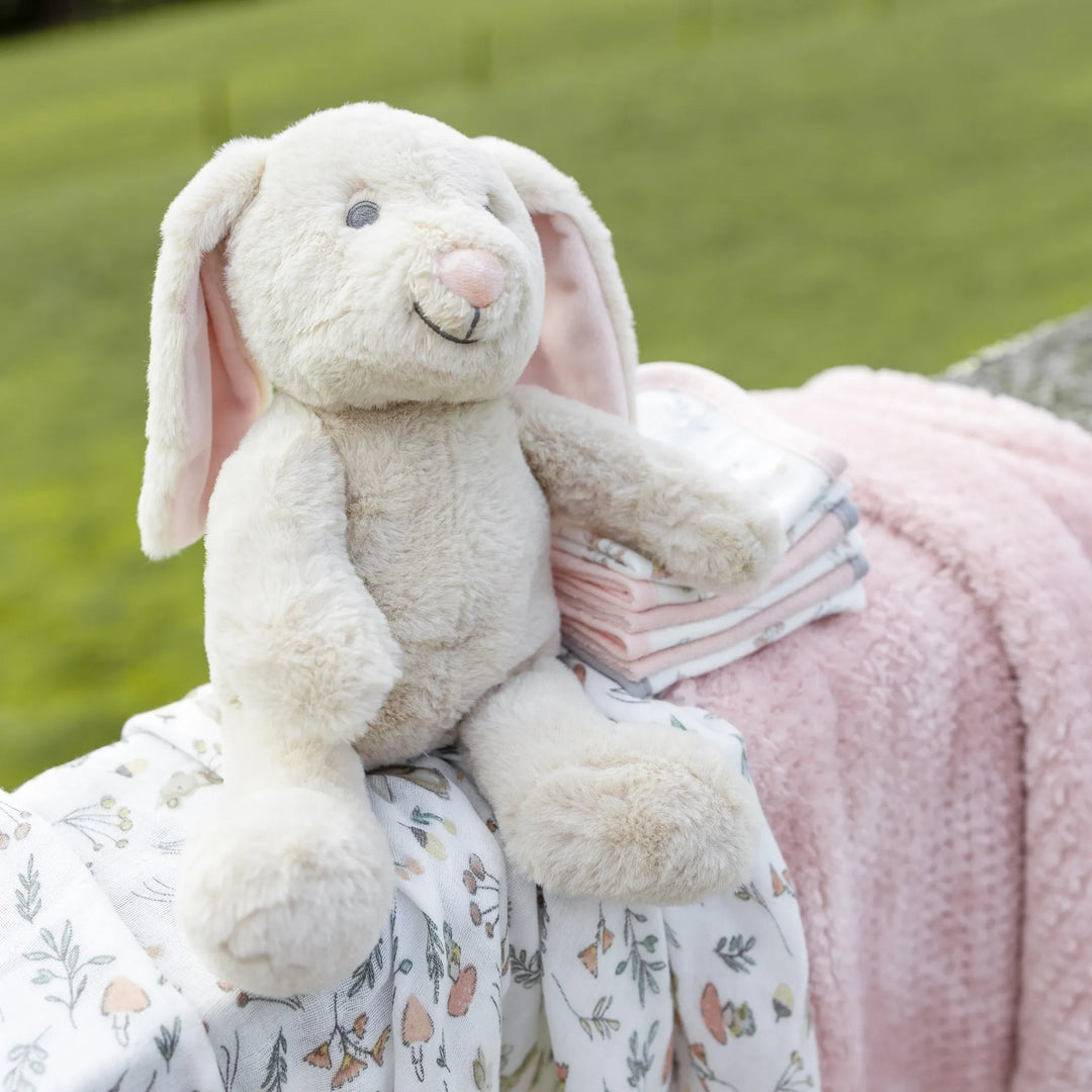 Plush bunny toy sitting on a floral blanket with a pink blanket underneath, against a green grass background.