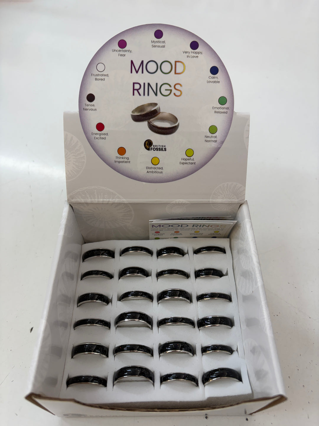 An open display box of mood rings showing multiple black metal bands with silver inner rings, with a circular insert card titled 'MOOD RINGS' above them on a white surface.