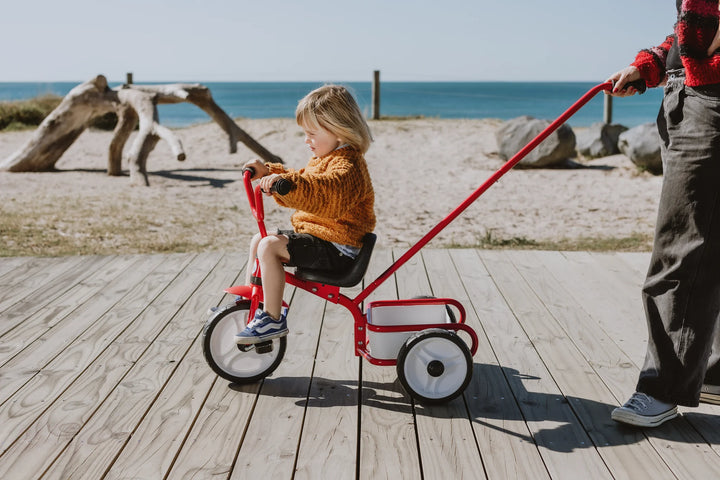 Child riding a red tricycle with an adult pulling it on a wooden boardwalk by the beach.