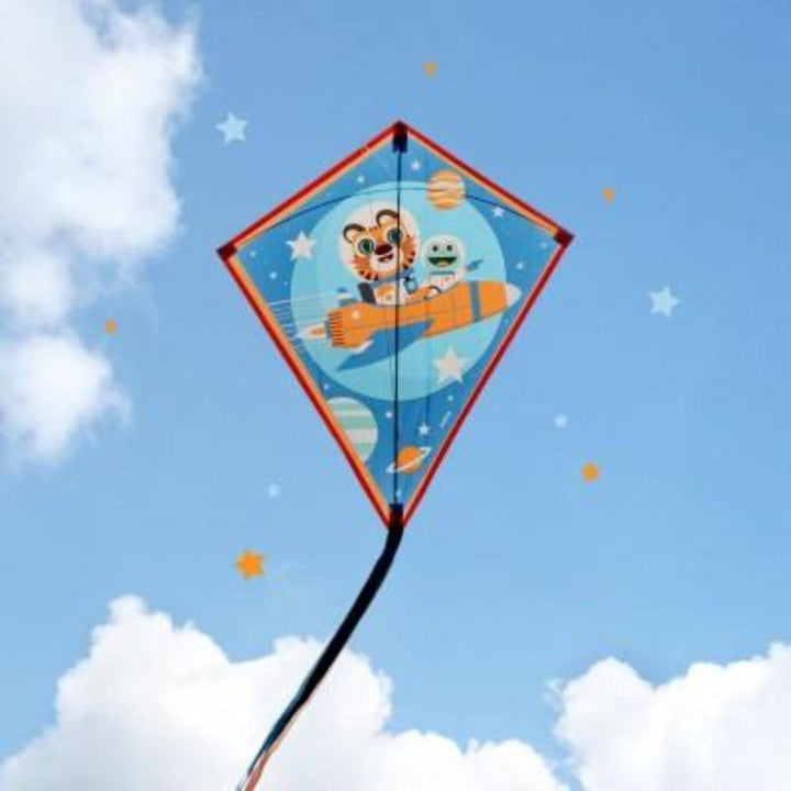 A diamond-shaped kite flying in a blue sky with white clouds. The kite features cartoon characters, including a tiger and a green creature, inside a blue space-themed design with stars and planets.