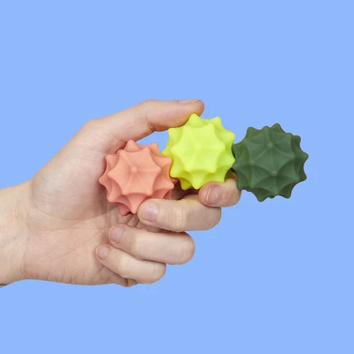 Hand holding three textured stress balls in orange, yellow, and green against a blue background