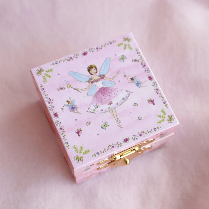A small pink square jewelry box decorated with a fairy illustration, floral border, and gold clasp, photographed on a soft pink background.