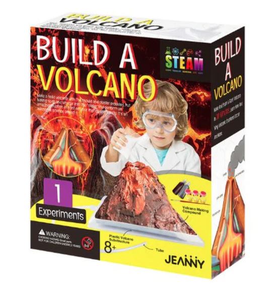 Box packaging for a children's science experiment kit titled 'Build a Volcano' by Jeanný. The front shows fiery graphics, a child in a lab coat holding a volcano model, and text indicating 1 experiment, 8+, and STEAM.