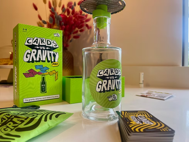 Cards vs Gravity Pro game set on a tabletop, featuring a clear glass bottle with a green cap and label, a bright green product box, cards, a pouch, and a small container in a home-like setting.