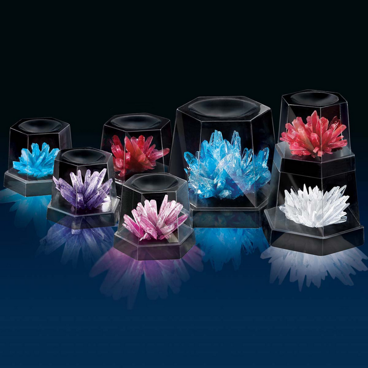 A set of eight black hexagonal crystal display boxes arranged on a dark reflective surface, each holding a different colored crystal cluster including blue, red, purple, pink, and white.
