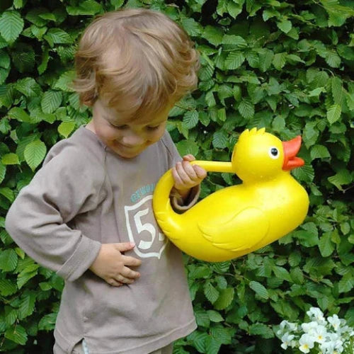 Child holding a large yellow rubber duck watering can in front of a green leafy background