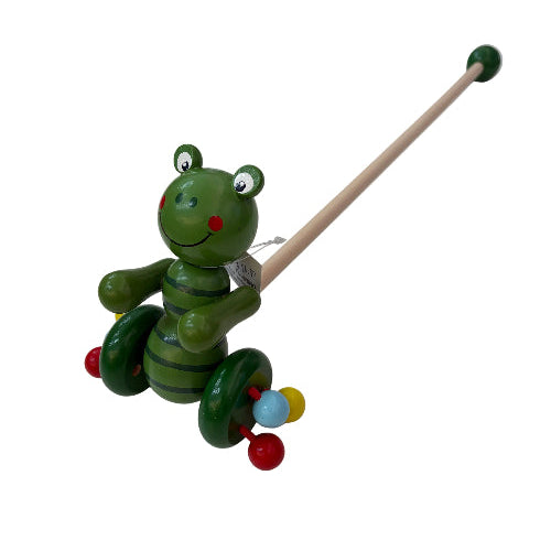 A green wooden frog toy with a long natural wood handle and colorful bead accents, photographed on a white tabletop next to a store display.