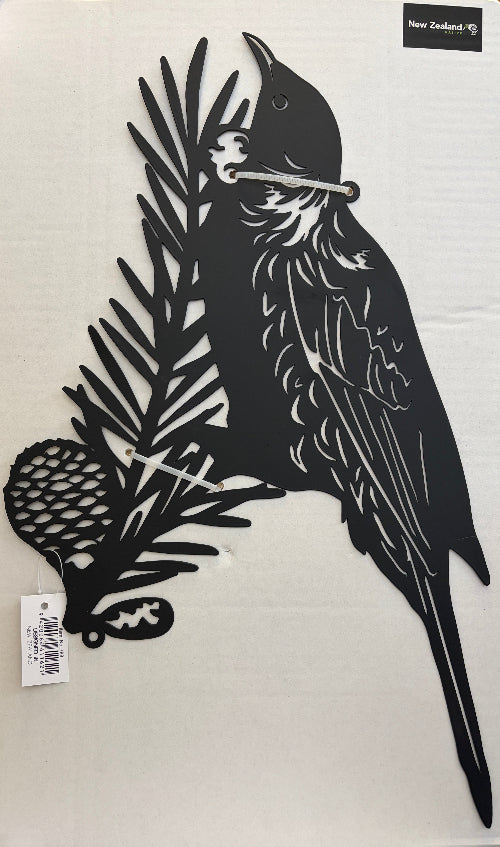 A black metal wall art piece featuring a tui bird perched on a pine branch with a pinecone, displayed on a white background with a small 'New Zealand' label and a barcode tag attached.