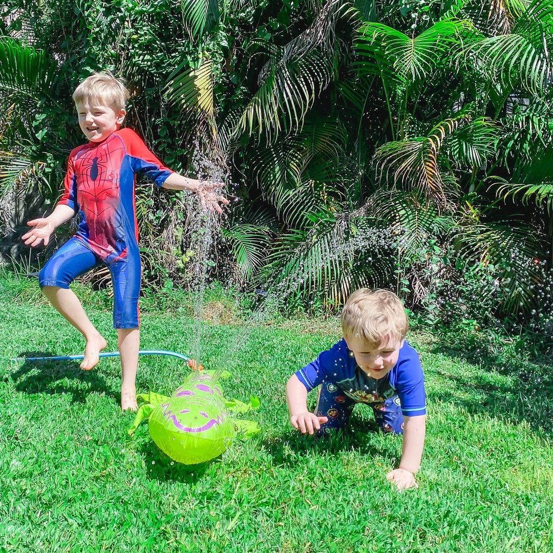 Two young boys playing outdoors on a grassy lawn with a sprinkler. The boy on the left wears a red and blue Spider-Man themed swim or play suit and is jumping in the water, while the boy on the right wears a blue long-sleeve shirt and crawls toward a bright green inflatable ball.