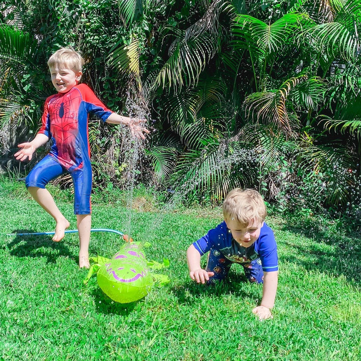 Two young boys playing outdoors on a grassy lawn with a sprinkler. The boy on the left wears a red and blue Spider-Man themed swim or play suit and is jumping in the water, while the boy on the right wears a blue long-sleeve shirt and crawls toward a bright green inflatable ball.