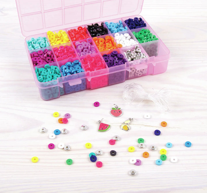 A pink plastic organizer box filled with many small colorful beads or charms, with several loose multicolored round beads and a few decorative charm pieces scattered in front on a light wood surface.