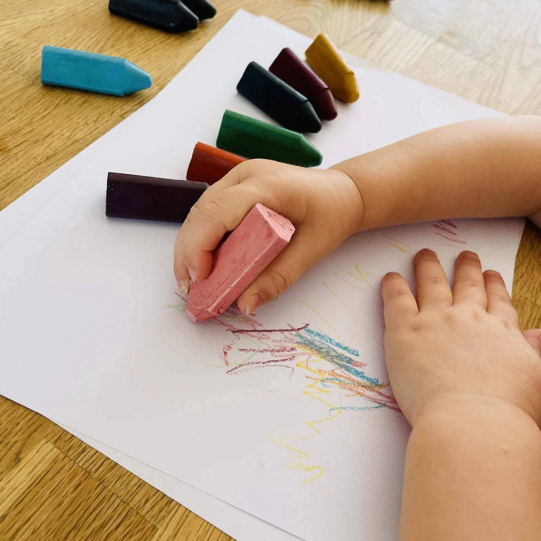 Child's hand holding a pink honeystick triangle crayon over a drawing on white paper with crayons scattered around.