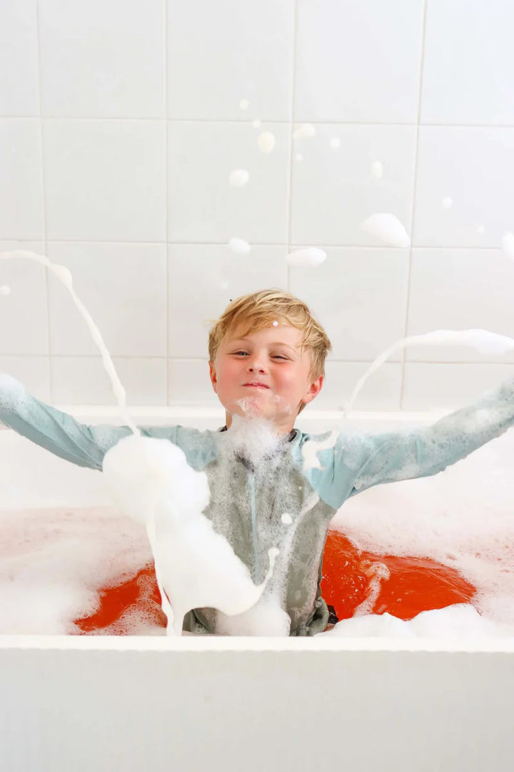 Child in a bathtub filled with bubbles and water, surrounded by white tiled walls.