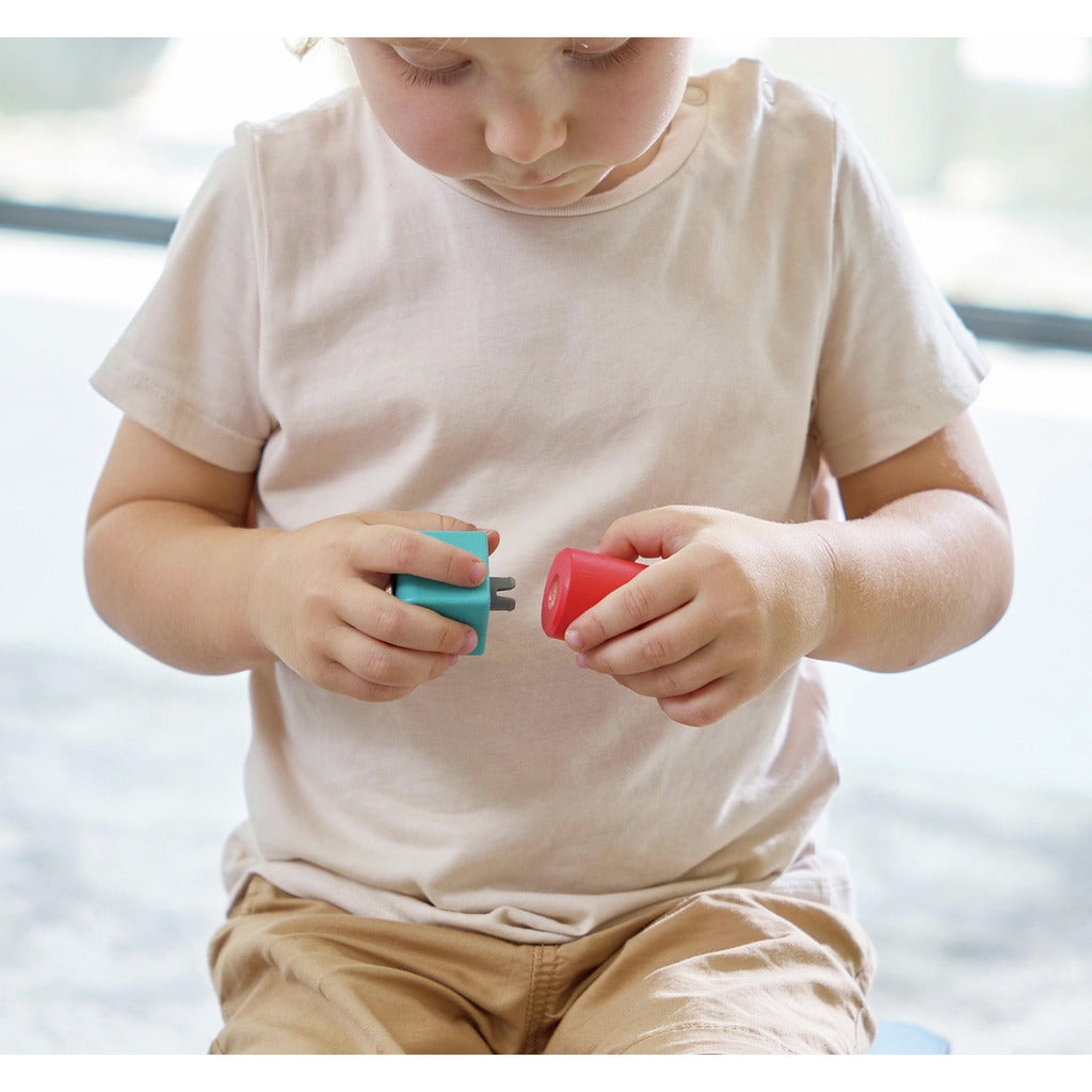 Child holding two small building toys, one blue and one red, against a blurred background. hape imagination blocks