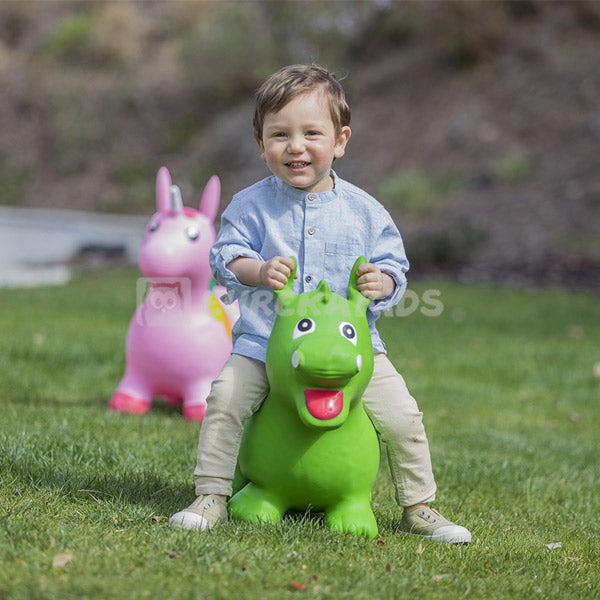 Child playing on a green inflatable dragon dinosaur in a grassy area with a pink unicorn toy in the background.