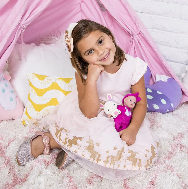 Young girl in a white dress with gold patterns, sitting on a pink cushion with adora tot doll, surrounded by a pink canopy.