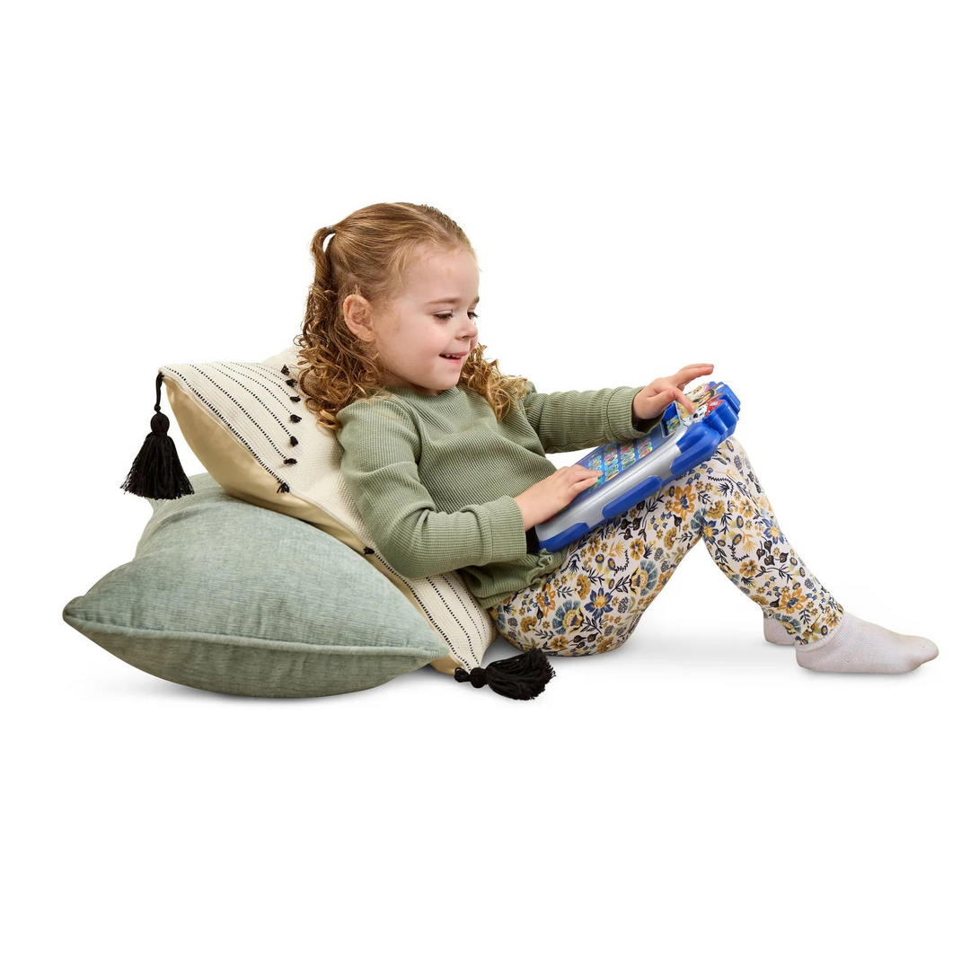 A young girl with curly hair sits on a green and cream decorative pillow, holding a blue and white tablet. She is wearing a sage green long-sleeve top and yellow, blue, and white patterned leggings against a solid white background.