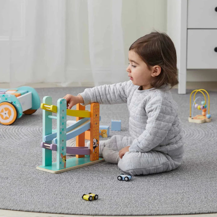 A toddler in a gray quilted pajama set sits on a gray carpet, playing with a colorful wooden multi-level race track toy. The room has soft white curtains and a white dresser in the background.