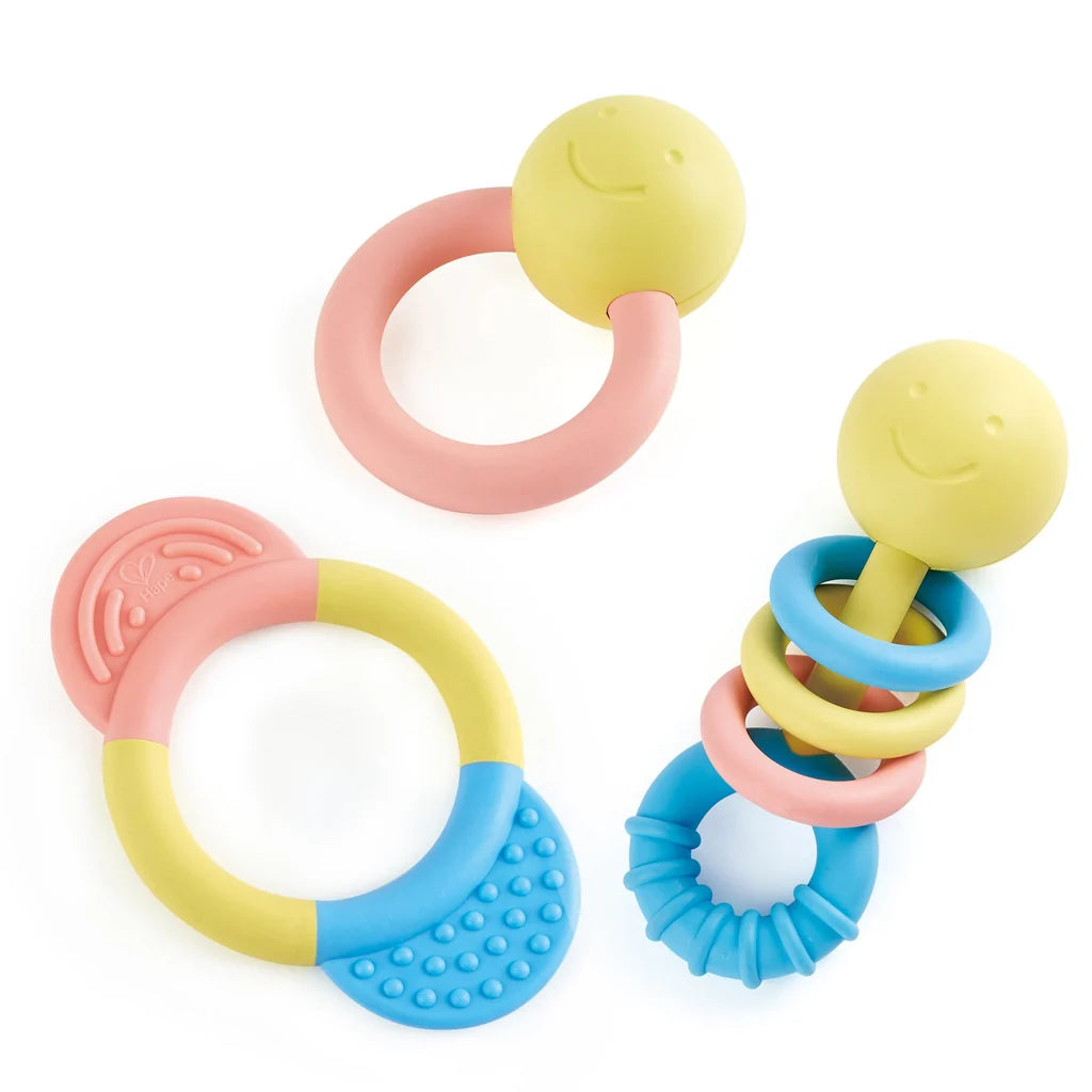 Three colorful baby teething toys on a white background: two ring teething toys with yellow smiley-face balls and one stacked ring teething toy with blue, yellow, pink, and blue rings.