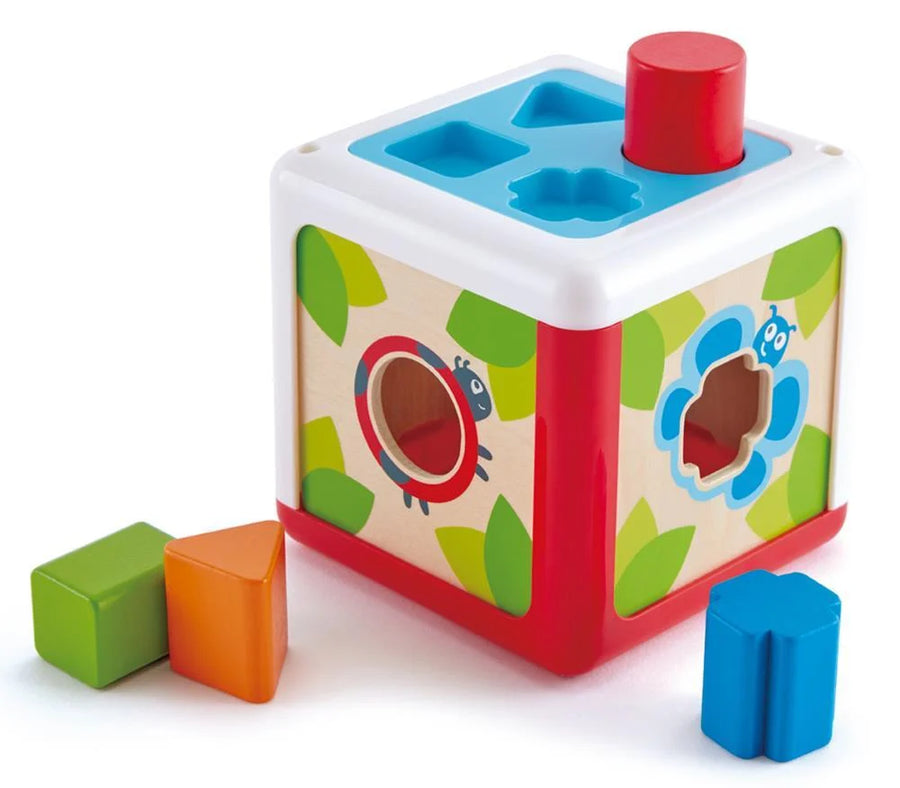 A colorful wooden shape-sorting cube toy with a red top cylinder, blue and green shape holes, and three loose blocks in green, orange, and blue on a white background.