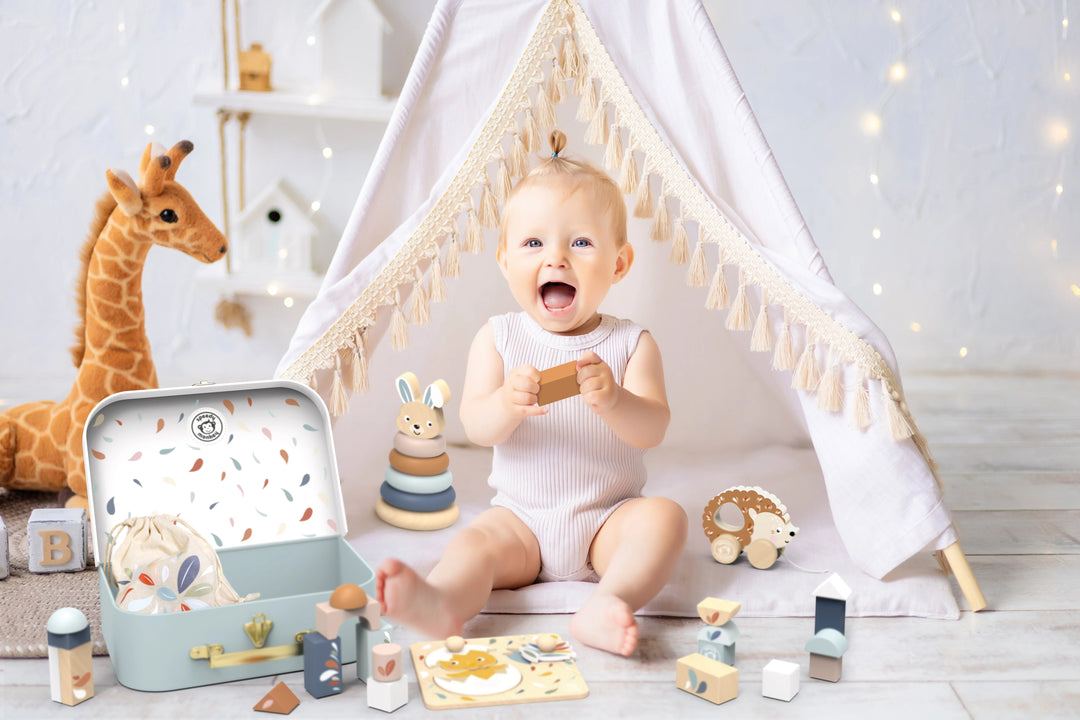 Baby sitting inside a small tent with toys around, including a suitcase and wooden toys.