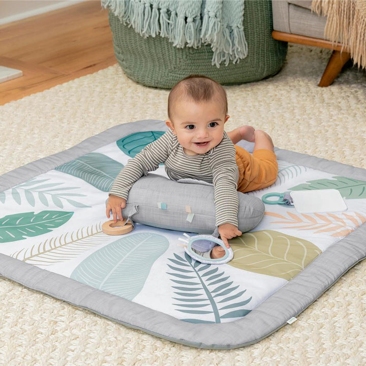 Baby playing on a leaf-patterned mat with toys on a carpeted floor.