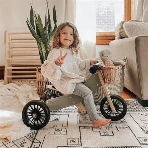 Child sitting on a kinder feet tiny toy tricycle in a cozy living room with a teddy bear.