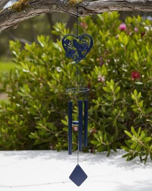 A tui metal wind chime hanging from a tree branch outdoors, with green shrubs in the background.