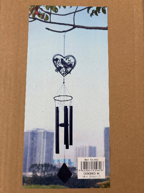 A decorative wind chime with a heart-shaped metal ornament featuring a tui bird design, hanging from a branch against a city skyline background. The chime has three black cylindrical tubes and a black diamond-shaped weight at the bottom.