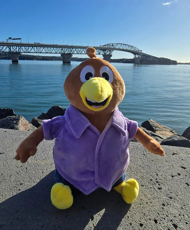 Plush toy with a purple shirt and yellow feet on a concrete surface by a waterfront with a bridge in the background.
