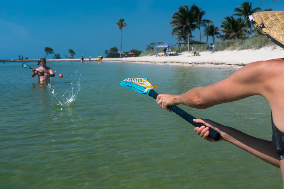 Two people playing with a Water Lacrosse Set in shallow water near a beach.