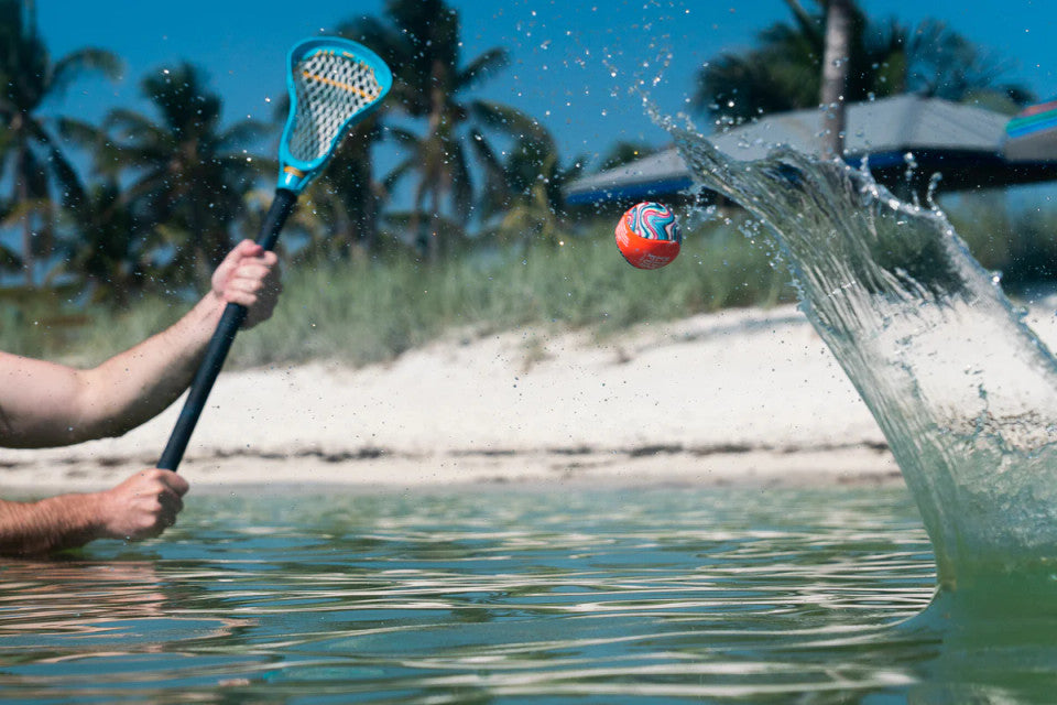 Person playing with a waboba lacrosse ball and stick in water near a beach