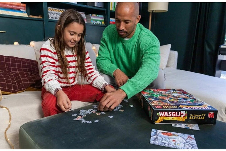 A man and a young girl are sitting on a couch in a living room, assembling a Wasgi Jigsaw puzzle together on a dark table. The puzzle box is visible on the right side of the table.