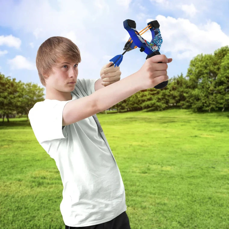 A young man in a white t-shirt aiming a blue and black slingshot outdoors in a grassy park under a cloudy sky.